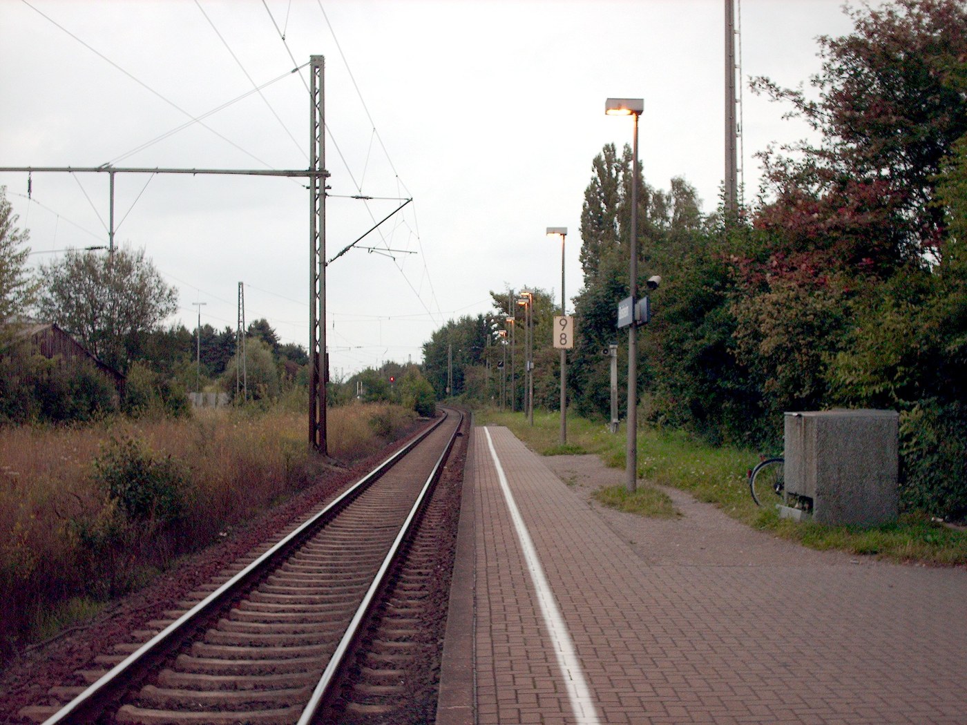 Bahnsteig und Gleise am Bahnhof Boisheim mit Oberleitungen, Bäumen und dem Bahnübergang im Hintergrund.