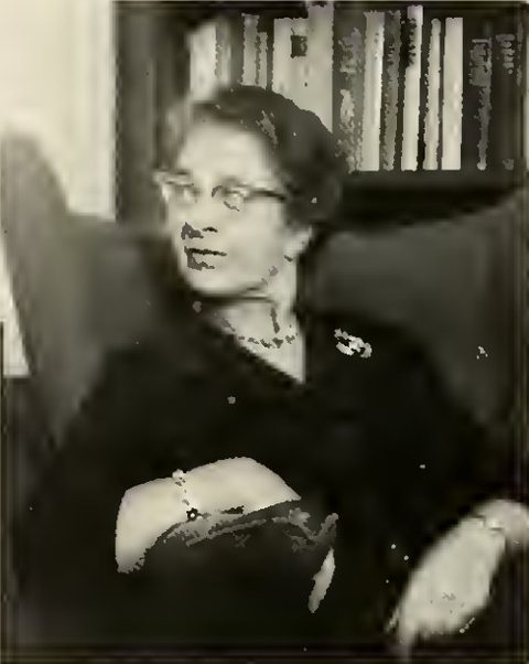 Hilde Bruch sitzt mit Brille und dunkler Kleidung vor einem Bücherregal, eine Hand auf dem Arm des Sessels.