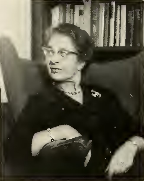 Hilde Bruch sitzt mit Brille und dunkler Kleidung vor einem Bücherregal, eine Hand auf dem Arm des Sessels.