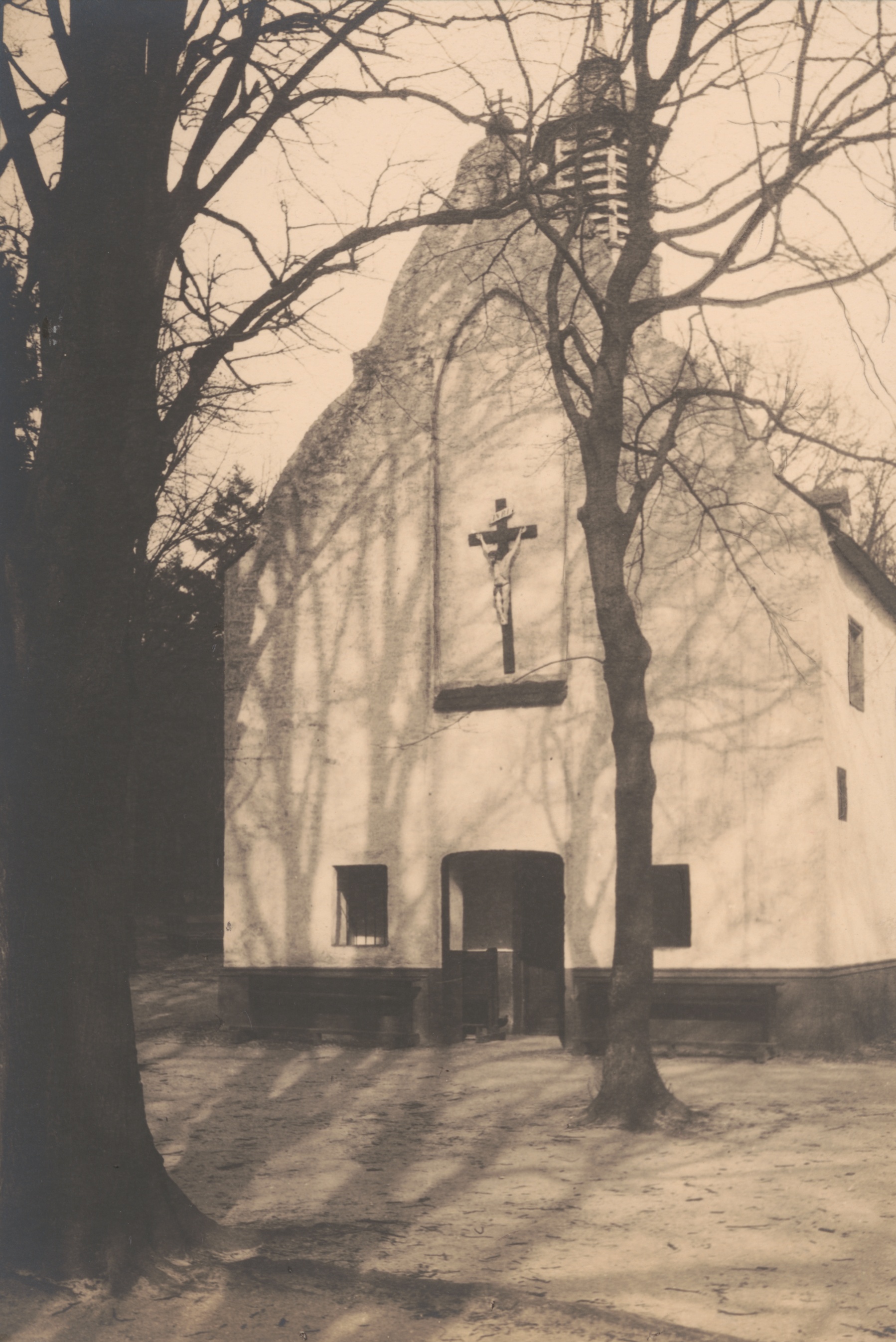 Sepiafarbene Originalfotografie der Giebelseite der Irmgardiskapelle zwischen kahlen Bäumen, mit Kreuz und offenem Eingang, 1911.