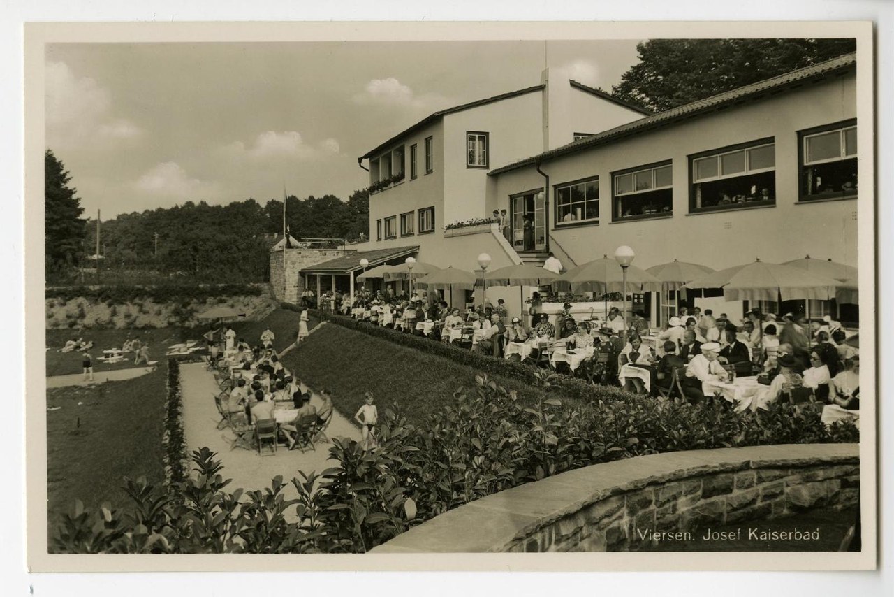 Terrasse des Josef-Kaiserbads mit besetzten Tischen und Sonnenschirmen vor dem Gebäude